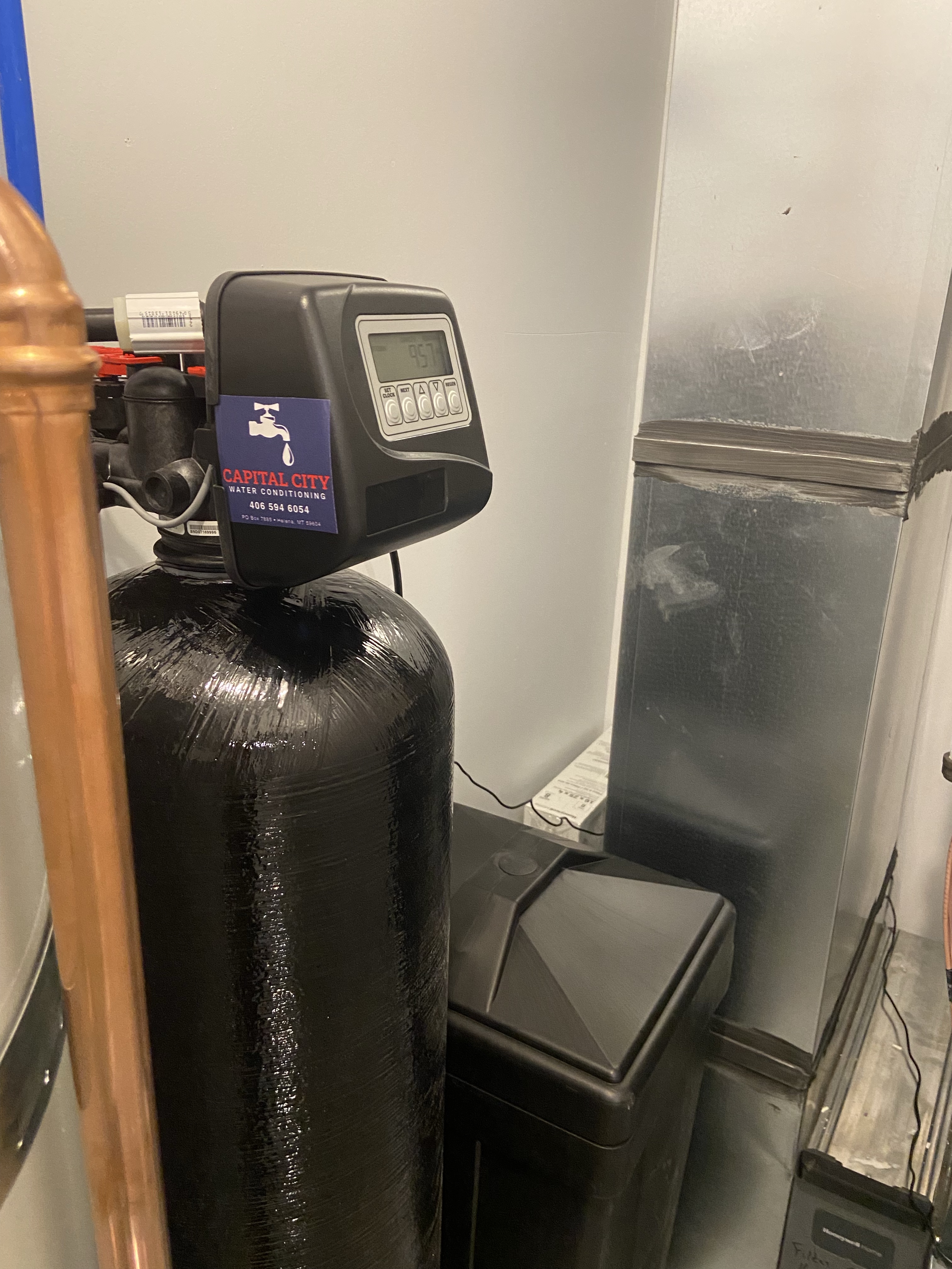 Black water softener tank next to an HVAC system. Sticker on side says Capital City Water Conditioning.